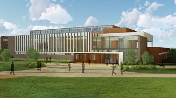 The exterior of the modern sports center at Strule Shared Education features large windows and is surrounded by lush grass and trees. People stroll along pathways in front of this vibrant education campus, creating a lively atmosphere.