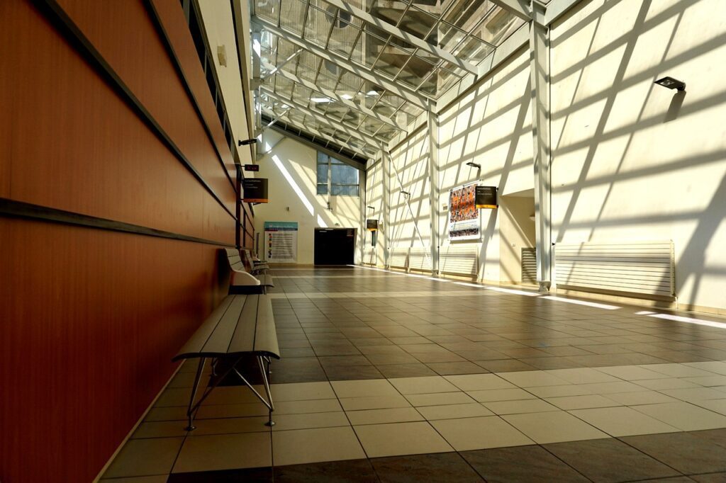 Spacious corridor with a tiled floor, benches on the left, and a glass ceiling casting shadows. A digital display highlights healthcare facilities information on the right wall.