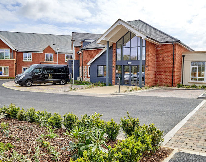 The exterior of the modern brick residential care home on Lace Hill features large windows. A black van is parked near the entrance, with landscaped greenery lining the paved driveway, creating a serene setting in this Buckingham neighborhood.
