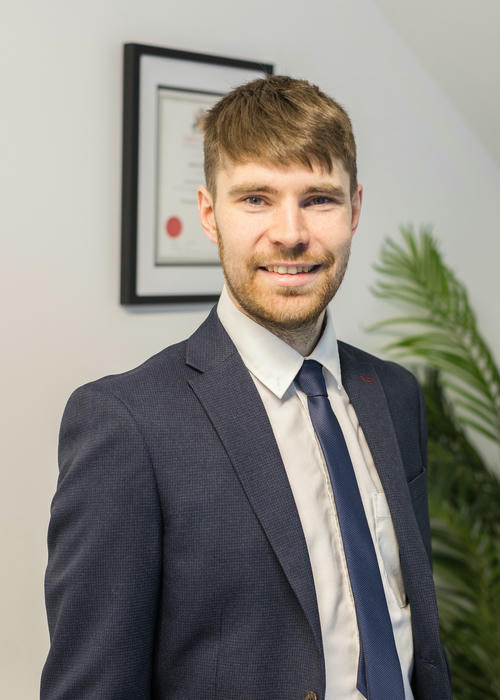 A man in a suit stands smiling in an office, proudly displaying a framed certificate for his expertise in fire risk assessment, with a lush plant adding a touch of greenery to the background.