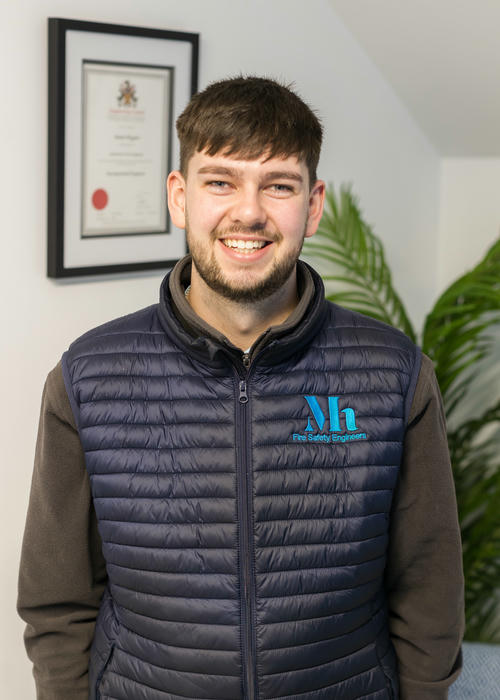 A person wearing a dark quilted vest with a logo stands indoors, smiling. In the background, alongside a framed certificate and a green plant, is evidence of their expertise in fire risk assessment.