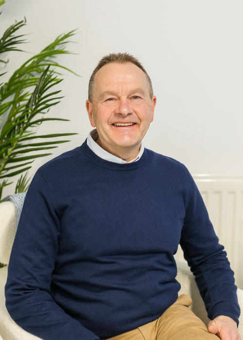 A smiling person sits comfortably in a chair, wearing a navy sweater and light shirt, while contemplating their recent fire risk assessment. A lush plant adds a touch of nature to the serene setting.