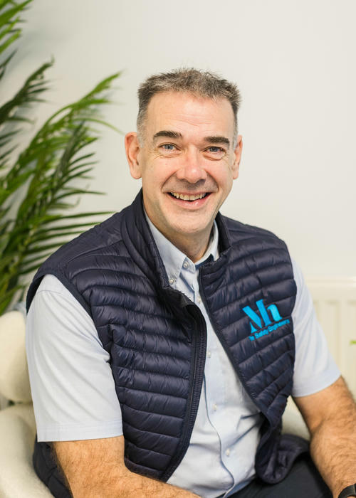A smiling man in a navy puffer vest with a logo sits indoors, surrounded by lush plants, as he reviews fire risk assessment notes.