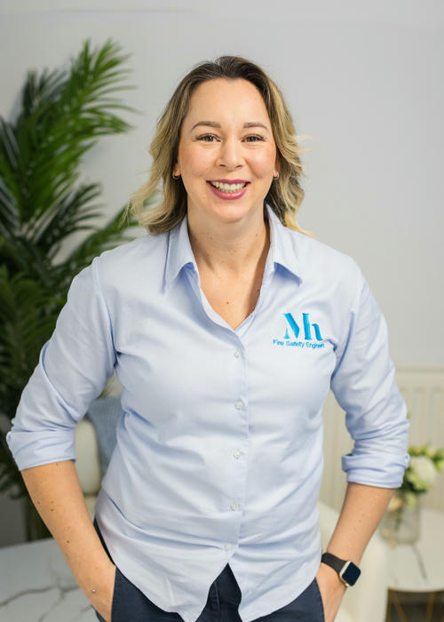 A smiling person in a light blue shirt with a logo stands confidently, hands on a table. Behind them, plants thrive against white walls, creating an inviting atmosphere. Their demeanor suggests expertise, perhaps even in areas like fire risk assessment.