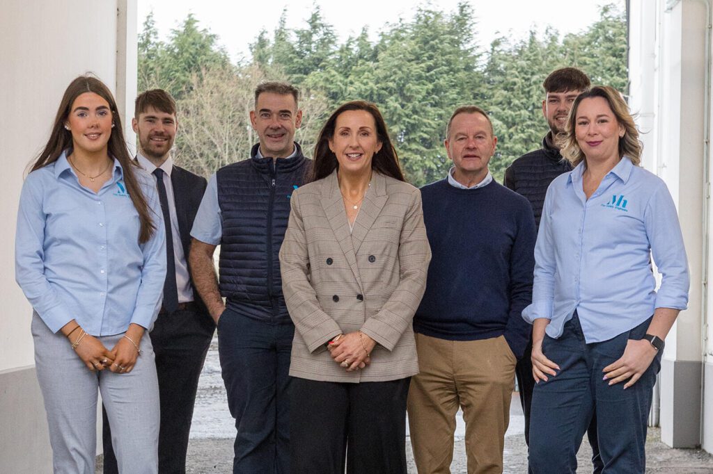 A group of seven people stands in a hallway, some in uniforms emblazoned with the company logo. Through the open door in the background, trees sway gently. They're gathered for a fire risk assessment to ensure safety and compliance.