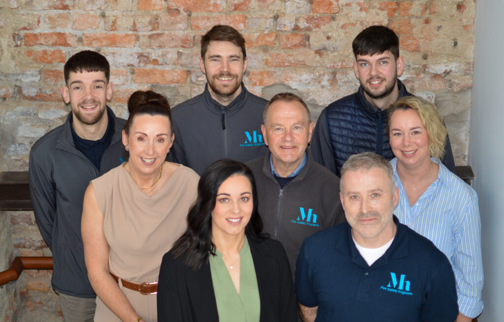 Eight people, five men and three women, stand and sit in two rows against a brick wall, wearing business casual and branded clothing, smiling at the camera after completing a fire risk assessment.