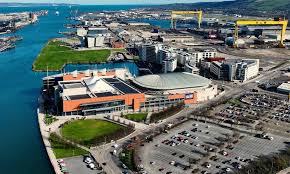 Aerial view of a waterfront city area with modern buildings, a large car park, green spaces, and two large yellow cranes near the water.