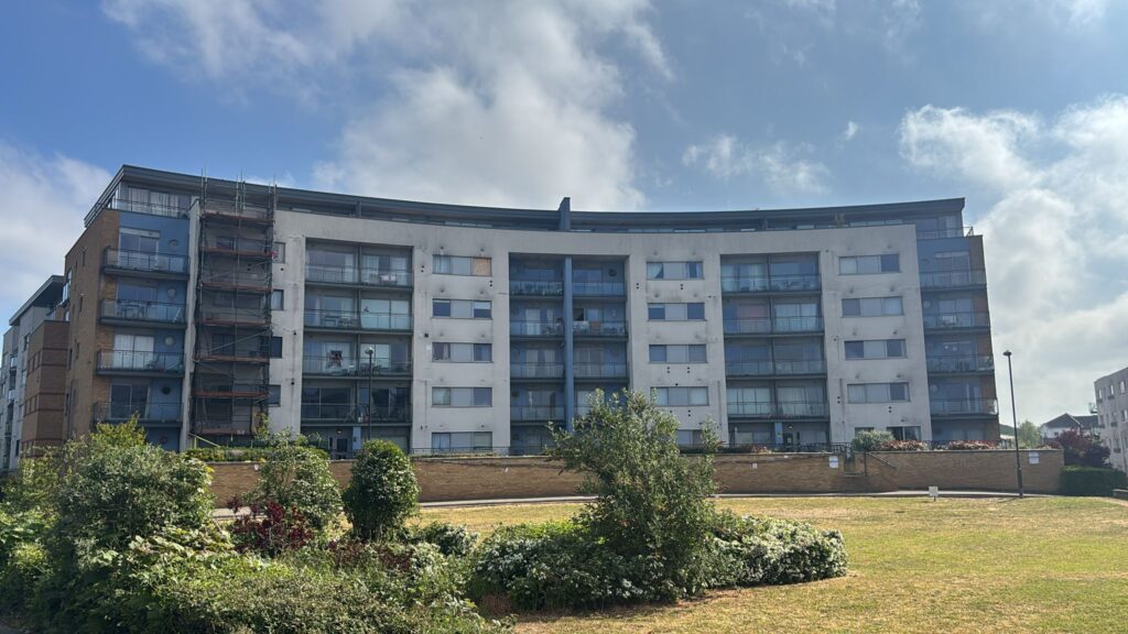 A modern five-story apartment building with balconies, glass panels, and some visible scaffolding stands behind a landscaped lawn under a partly cloudy sky.